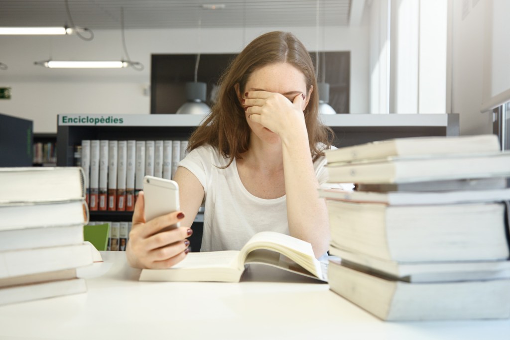 Étudiante assise à un bureau entouré de livres, tenant son téléphone d'une main et se tenant la tête de l'autre, l'air dépassée.