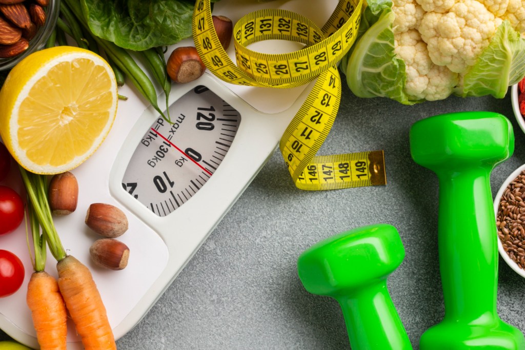 Pèse-personne recouvert de fruits frais, fruits secs et légumes, avec des haltères posés à côté.